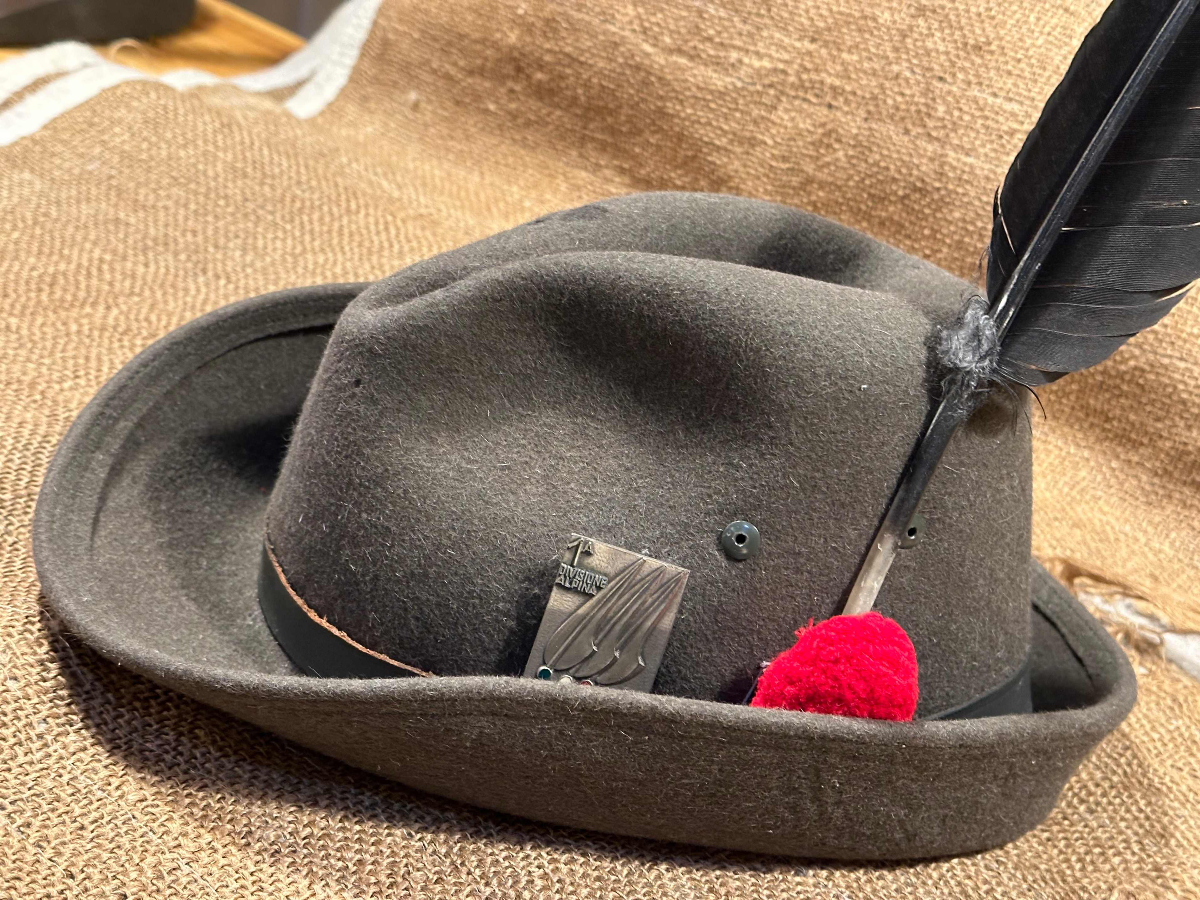WWII Italian Alpini mountain troops hat — grey-green felt with red pompom, eagle badge, and black raven feather